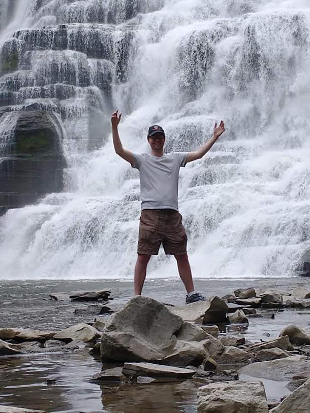 Gavin stands with his arms up in front of a waterfall.