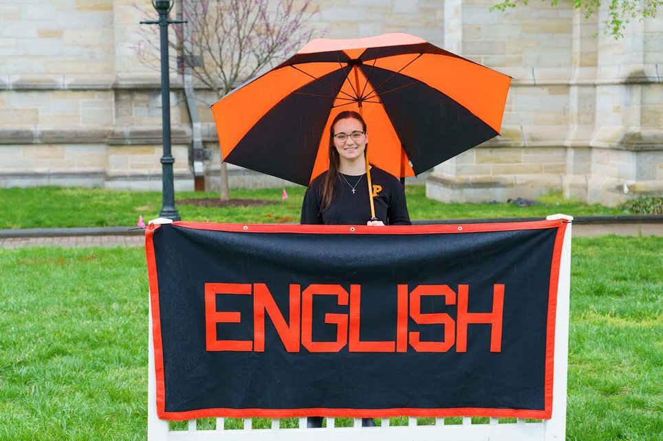 Selena smiles at camera. She stands under an orange and black umbrella and behind a banner reading "English."