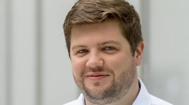 Head-and-shoulders portrait of a person with short light-brown hair and trimmed facial hair wearing a light blue button-down shirt, standing indoors against a softly blurred light gray wall.