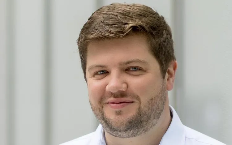 Head-and-shoulders portrait of a person with short light-brown hair and trimmed facial hair wearing a light blue button-down shirt, standing indoors against a softly blurred light gray wall.