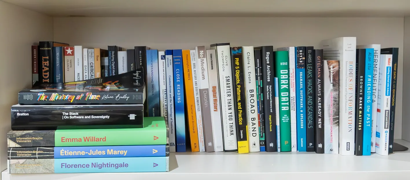 Wide bookshelf filled with assorted hardcover and paperback books, including titles on data, technology, history, and design, arranged upright and stacked horizontally.