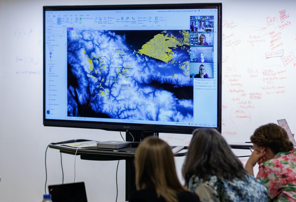 Three people look at a large screen where people are presenting on Zoom