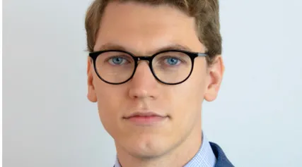 Head-and-shoulders portrait of a person wearing round black glasses, a blue suit jacket, a patterned tie, and a pocket square, standing against a plain light gray background.