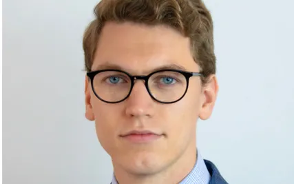 Head-and-shoulders portrait of a person wearing round black glasses, a blue suit jacket, a patterned tie, and a pocket square, standing against a plain light gray background.
