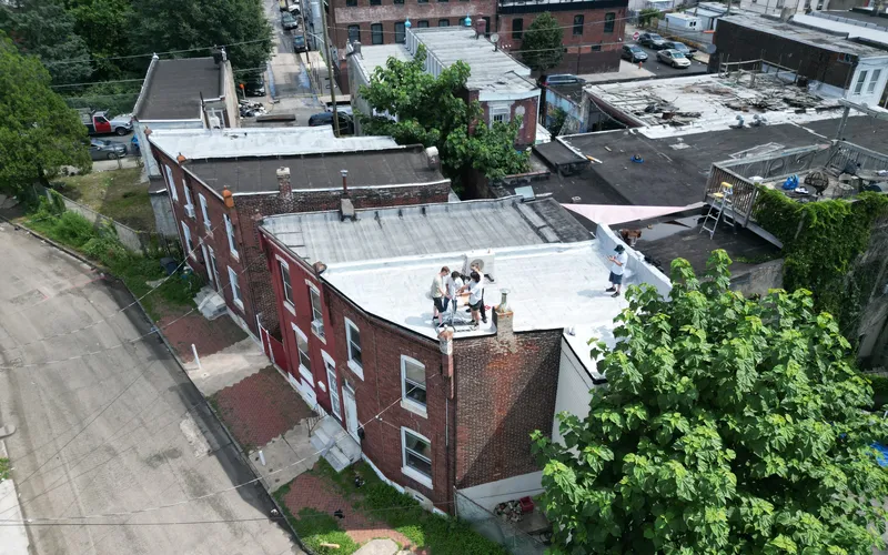Aerial view of a city block with brick row houses and flat rooftops, including several people gathered on one rooftop and a deck with furniture on another, surrounded by trees and parked cars along narrow streets.