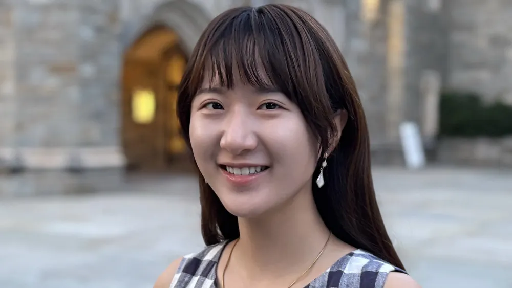 Hao_Tan_Bio_Photo2Head-and-shoulders outdoor portrait of a person standing in front of a stone building with an arched entrance, wearing a sleeveless checkered top and a necklace.