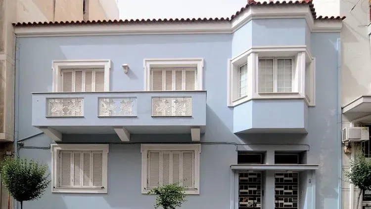 Front view of a light blue two-story house with white shuttered windows, a small balcony, and a protruding bay window, flanked by neighboring buildings.