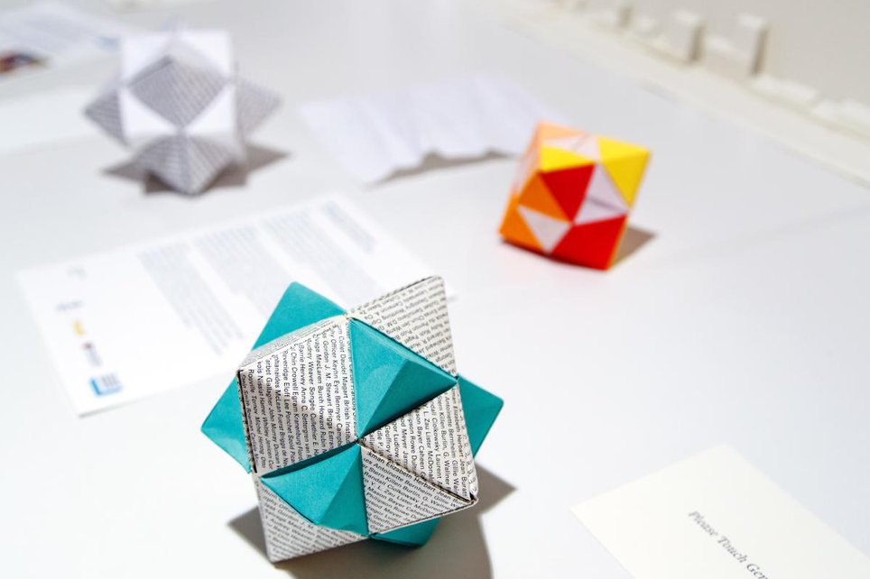 Three pieces of origami sit on a table. The front one is made from a blue-green cube and a white octahedron. The octahedron is printed with names.