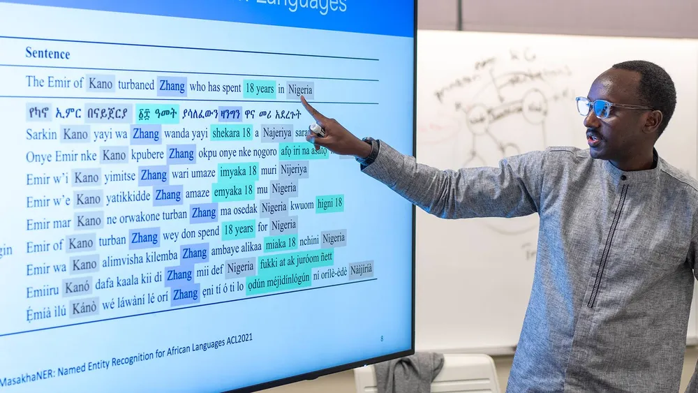 Person gesturing toward a large display showing a multilingual sentence comparison and named-entity recognition examples for African languages, with a whiteboard in the background.