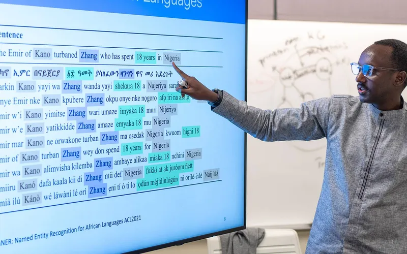 Person gesturing toward a large display showing a multilingual sentence comparison and named-entity recognition examples for African languages, with a whiteboard in the background.