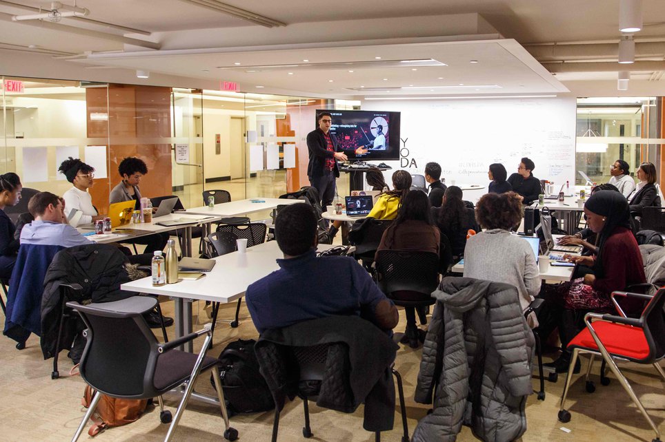 A man gestures towards a large TV with visualizations in front of a seated group of students with laptops.