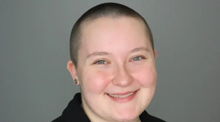 Studio portrait of a person with a shaved head wearing a dark jacket over a patterned shirt, a necklace with a crescent pendant, and small earrings against a plain gray background.