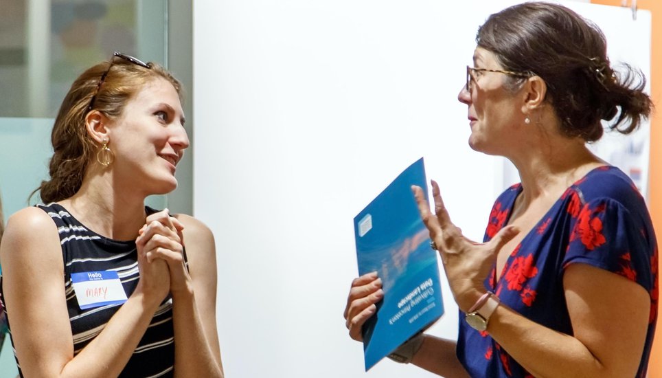 Two women having an animated conversation; one holds a blue folder.