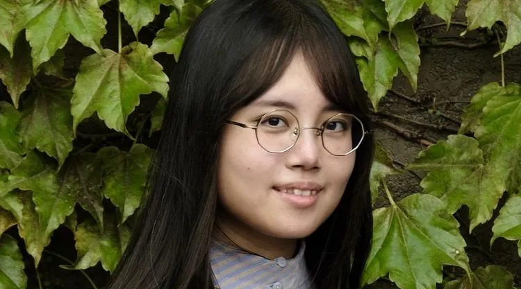 Head-and-shoulders photo of a person with long dark hair and round glasses wearing a striped button-up shirt, standing in front of a dense wall of green ivy leaves.