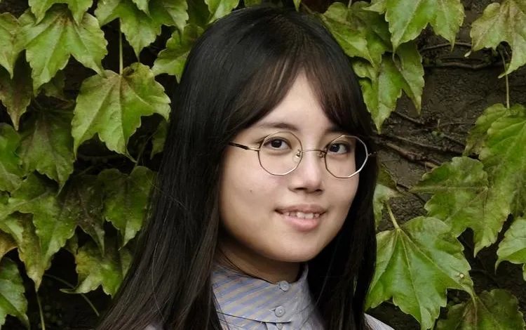 Head-and-shoulders photo of a person with long dark hair and round glasses wearing a striped button-up shirt, standing in front of a dense wall of green ivy leaves.