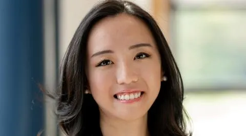 Studio-style head-and-shoulders photo of a person with shoulder-length dark hair wearing a sleeveless light top, standing indoors against a softly blurred background with a window and blue panel.