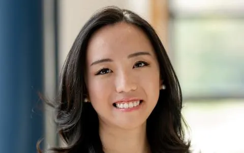 Studio-style head-and-shoulders photo of a person with shoulder-length dark hair wearing a sleeveless light top, standing indoors against a softly blurred background with a window and blue panel.