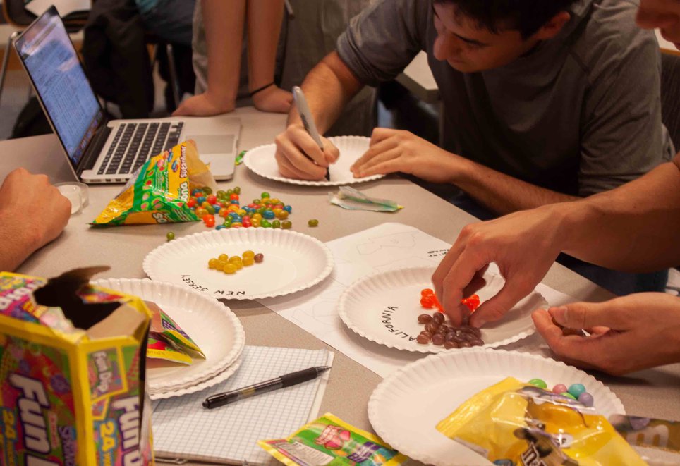 Students arrange jelly beans onto paper plates on a table filled with candy and condiments.