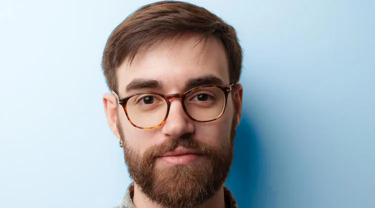 Head-and-shoulders portrait of a person with short brown hair, a full beard, and round glasses, wearing a patterned button-up shirt against a solid light blue background.