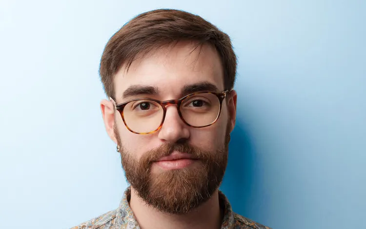 Head-and-shoulders portrait of a person with short brown hair, a full beard, and round glasses, wearing a patterned button-up shirt against a solid light blue background.