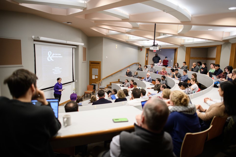 A woman in a purple shirt stands in front of a projector screen in a large lecture hall filled with attendees.