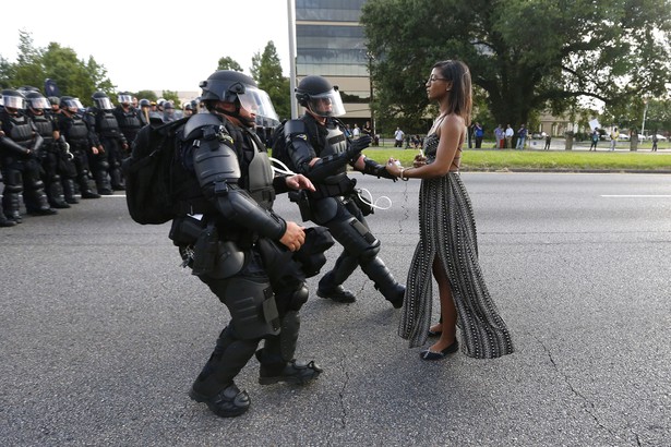 On the left, two law enforcement officers walk forward wearing protective equipment. On the right, a protester stands tall, facing the officers.